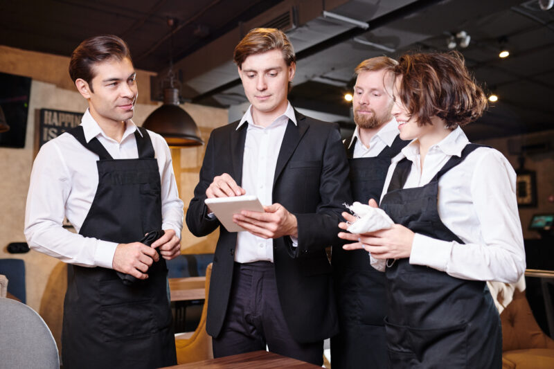 Serious restaurant manager dividing responsibilities among waite Serious confident handsome young restaurant manager using tablet while dividing responsibilities among waiters in modern establishment