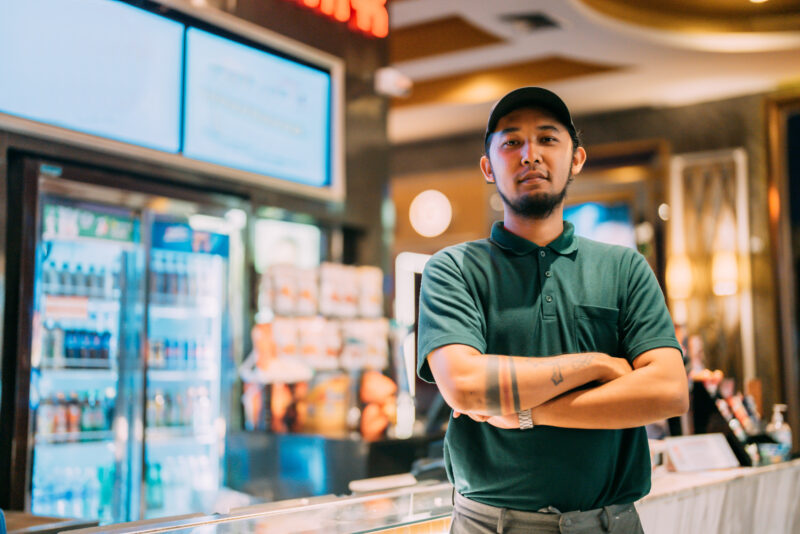 Portrait of asian tattooed cinema staff man against popcorn counter. He’s wearing a uniform. Portrait of tattooed cinema staff man against popcorn counter. He's wearing a uniform.