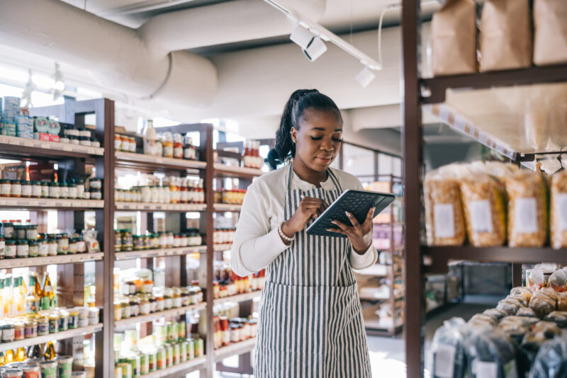 African employee at the organic convenience store uses a digital tablet for inventory, African employee seamlessly integrates technology into inventory management, using a digital tablet to track and organize items
