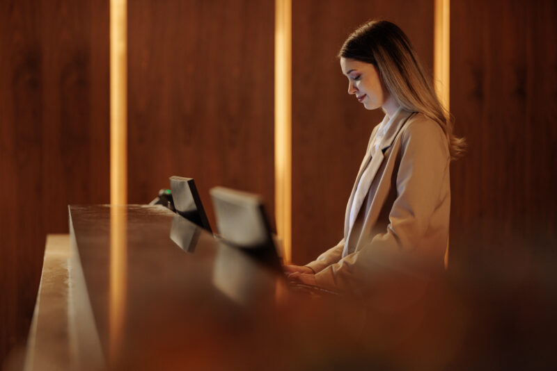 Hotel receptionist working at computer in lobby Young woman working at a hotel reception desk, engaging with guests while skillfully using a computer in a modern, cozy lobby setting