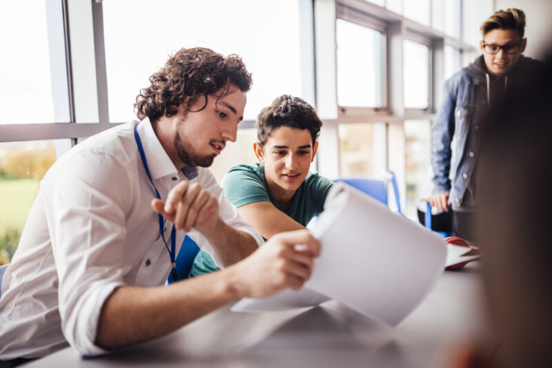Great School Work Teacher showing a student some results on his paperwork. The student looks happy as they sit side by side at a table. Another student looks on.