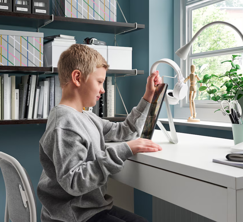 Headset-Stand A boy sits at a desk removing a pair of headphones from a white metal stand.