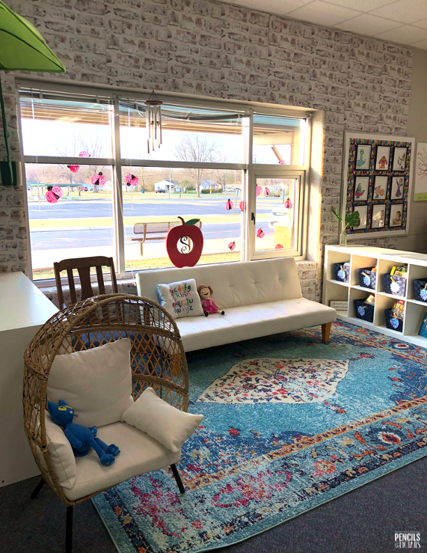A classroom setting is seen. There is a white chair, a futon, a large area rug, and a bookshelf with cubbies with baskets in it.