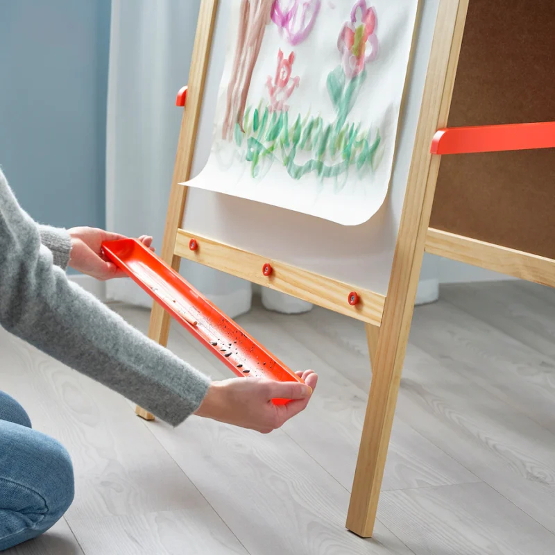 Mala-Easel An easel is shown with a painting of lowers on it. Hands are shown removing a basin from the bottom of the easel in this example of Ikea classroom.