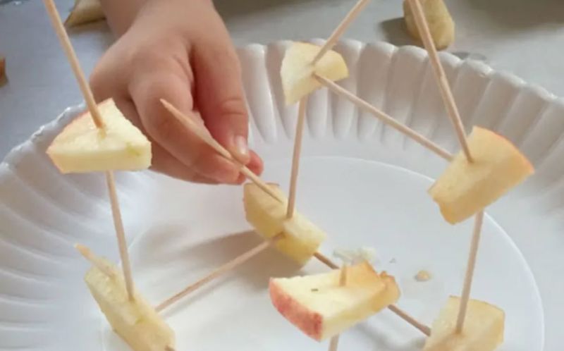 Apple Toothpick Tower STEM Challenge Child using toothpicks and pieces of apple to build a tower on a paper plate as an example of science activities for preschoolers