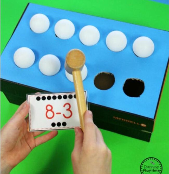 Dollar Store Hacks: Ping Pong Subtractions Student using a wooden mallet to tap ping pong balls into a cardboard box while holding a card saying 8-3