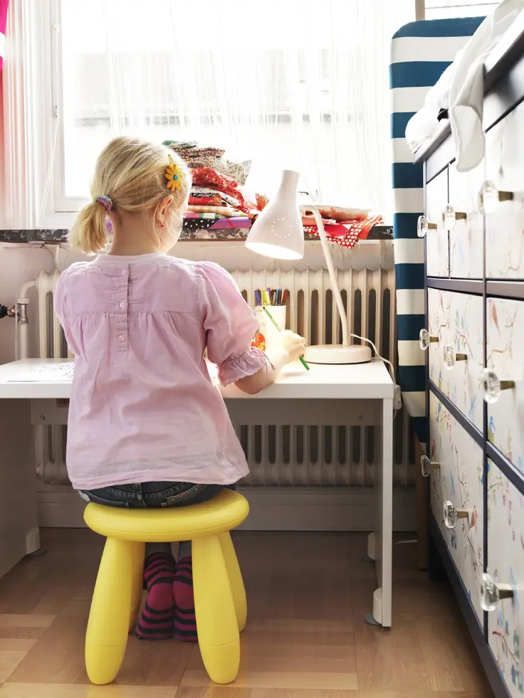 A little girl sits on a yellow, plastic stool at a desk.