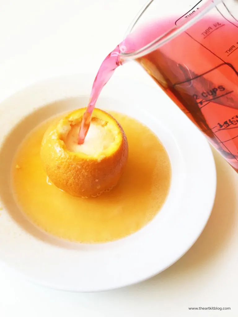 Orange Baking Soda Volcano An orange is shown in a bowl with a pitcher of red liquid pouring into the cut off top of it.