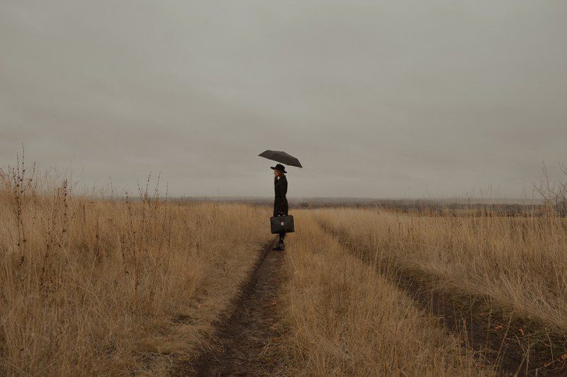 Picture Writing Prompts: Mary Poppins Woman standing in the middle of a wheat field on a gray day, holding an umbrella and bag