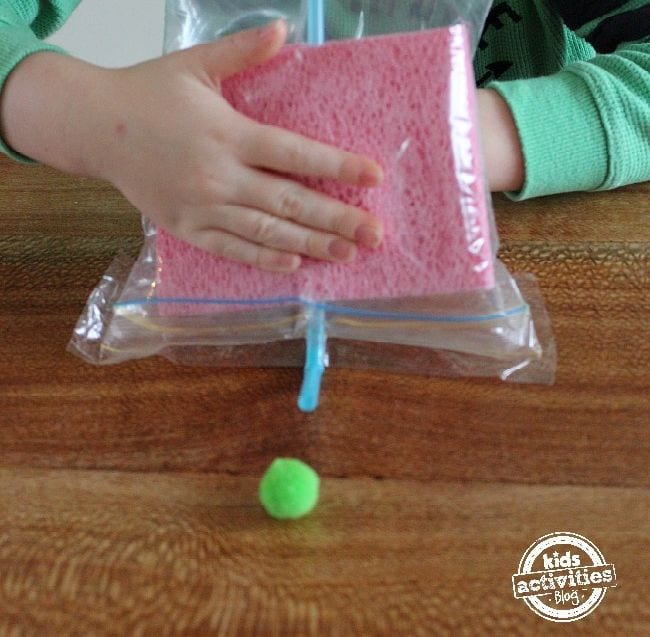 preschool-science-air-pressure Young student using a straw in a plastic bag with sponges to move a green pompom across a wood table