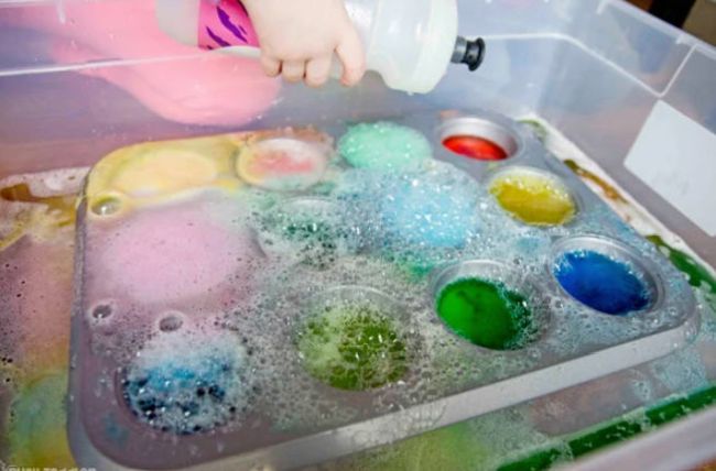 rainbow-baking-soda-and-vinegar Preschool student squeezing a bottle of vinegar into muffin tin cups filled with baking soda and food coloring as an example of science activities for preschoolers
