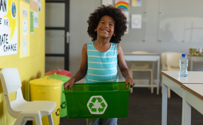 Smiling african american elementary schoolboy holding container with recycle symbol. unaltered, education, childhood, learning, environmental cleanup, sustainable lifestyle and school concept.