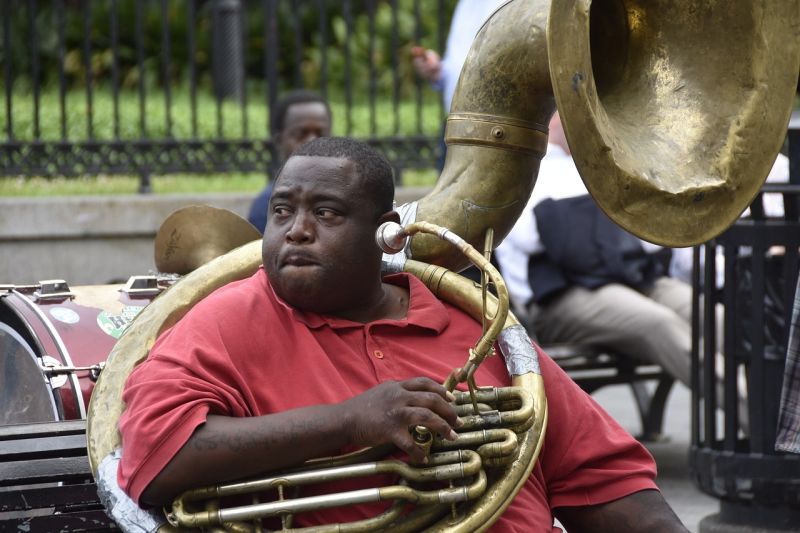 Picture Prompt: Sousaphone Player Black man in a red shirt sitting on a bench, wearing a sousaphone