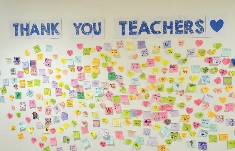 Teacher Appreciation Week Wall A wall has large signs across the top reading Thank You Teachers. Underneath are colorful sticky notes with messages of teacher appreciation written on them.