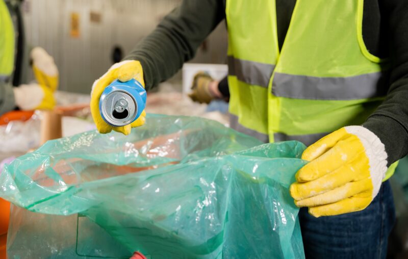 Cropped view of man in protective vest and gloves putting tin can in plastic bag while working near